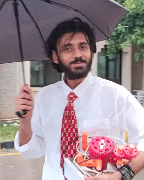 Taimoor Mazhar in the rain, holding an umbrella and a red 3D-printed gear model.