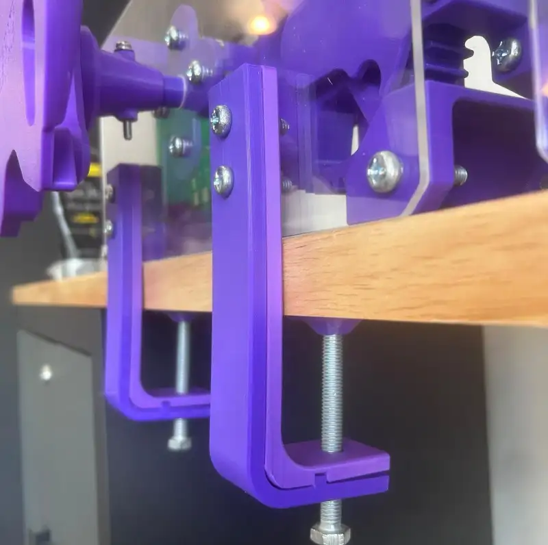 A purple printed clamp tightened under a desk with a threaded rod and knob.